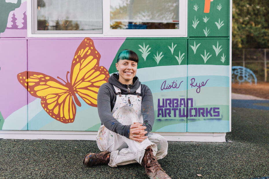 Artist Aistė Rye sitting in front of a mural she designed for the Northwest School for Deaf and Hard-of-Hearing Children. she is sitting in front of a butterfly her artist's signature and the Urban ArtWorks tag.